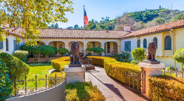 A sunny courtyard at Foothill Retirement featuring a pathway lined with trimmed hedges leading to a building with arched doorways and windows. Two lion statues stand on pedestals at the entrance of the pathway, and an American flag is visible in the background with hills and trees surrounding the area.