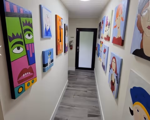 A narrow hallway with light-colored walls and wood-patterned flooring, decorated with colorful modern art paintings on both sides. At the end of the hallway is a black-framed door with a frosted glass panel and a fire extinguisher mounted on the wall to the left.