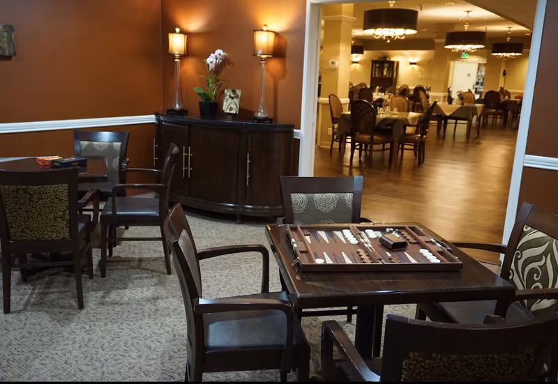 A cozy indoor seating area with several wooden tables and chairs. One table in the foreground has a backgammon board set up. The room has warm brown walls, carpeted flooring, and two tall lamps on a dark wooden cabinet with a potted plant and a framed picture. In the background, there is a dining area with multiple tables and chairs under hanging light fixtures.