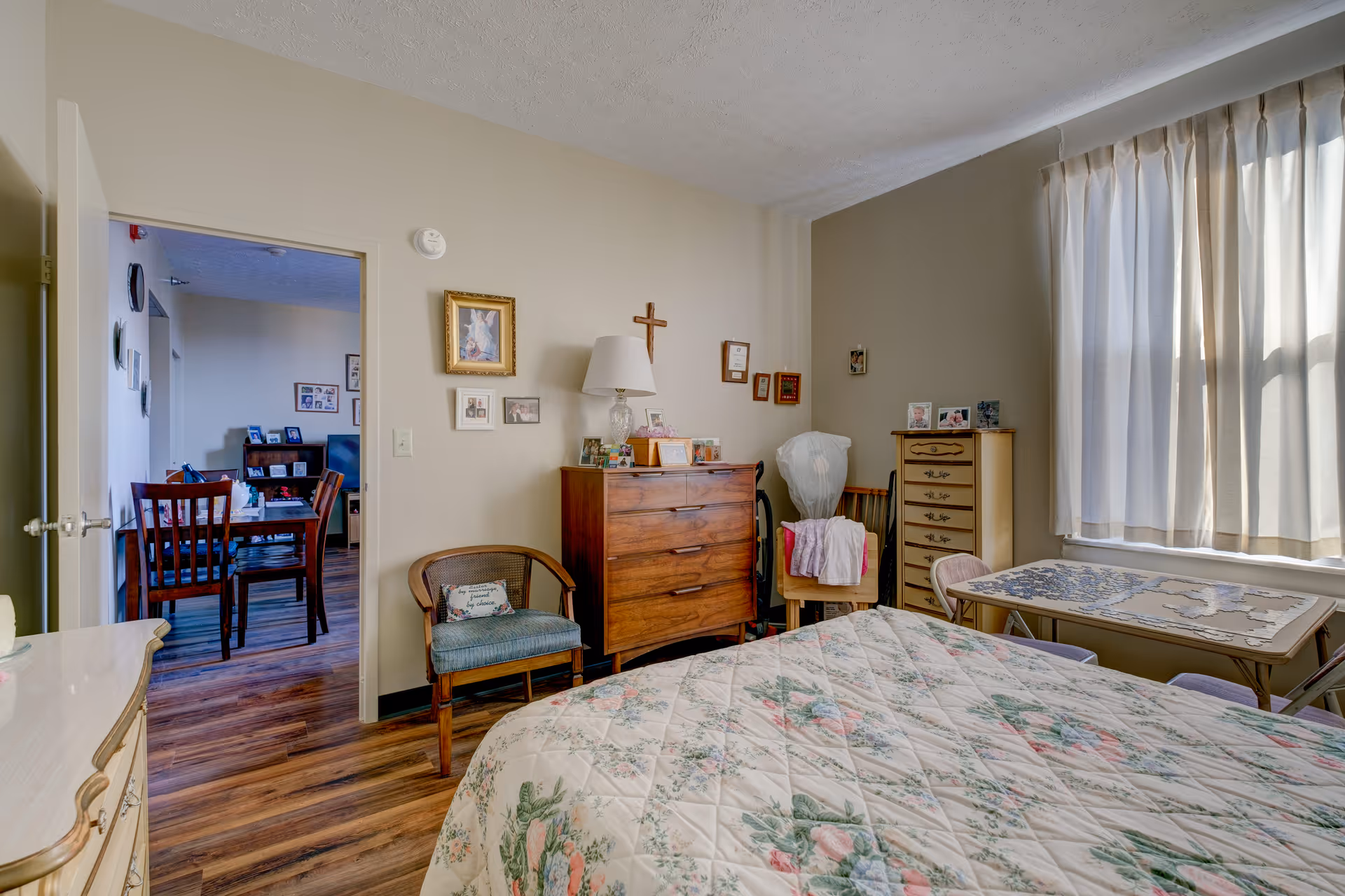 A cozy bedroom with a floral patterned bedspread on the bed, a wooden dresser with a lamp and framed pictures on top, a chair with a cushion, and a small table with a puzzle in progress near a window with light curtains. The door is open, showing a dining area with a wooden table and chairs.