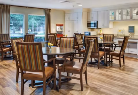 Dining room with round tables, striped-upholstered chairs, wood floors, and a kitchenette in the background.