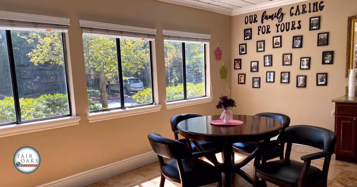 A bright room with three large windows showing greenery outside. Inside, there is a round wooden table with four black chairs around it. A small vase with purple flowers sits on a pink placemat in the center of the table. On the wall, there are framed photos arranged under the text 'OUR family CARING FOR YOURS'. A countertop with a paper towel roll is visible on the right side. The Fair Oaks Estates logo is in the bottom-left corner.