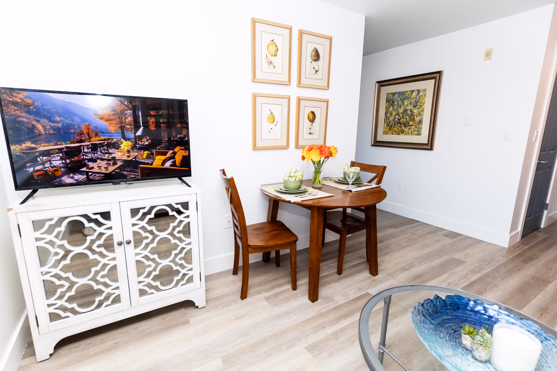 A small dining area with a round wooden table set for two with green plates, glasses, and a vase of orange flowers. Next to the table is a white cabinet with a decorative lattice design, holding a flat-screen TV displaying an outdoor scene with a lake and autumn trees. The walls are white and decorated with framed artwork, and the floor is light wood.
