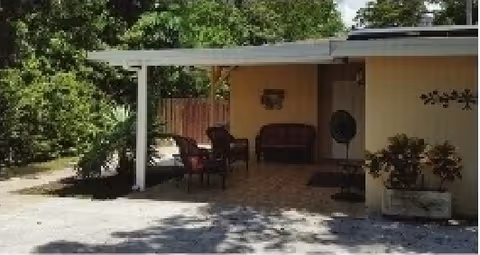 Covered outdoor patio area attached to a yellow building with seating including chairs and a bench. The patio is shaded by a white roof and surrounded by greenery and trees.