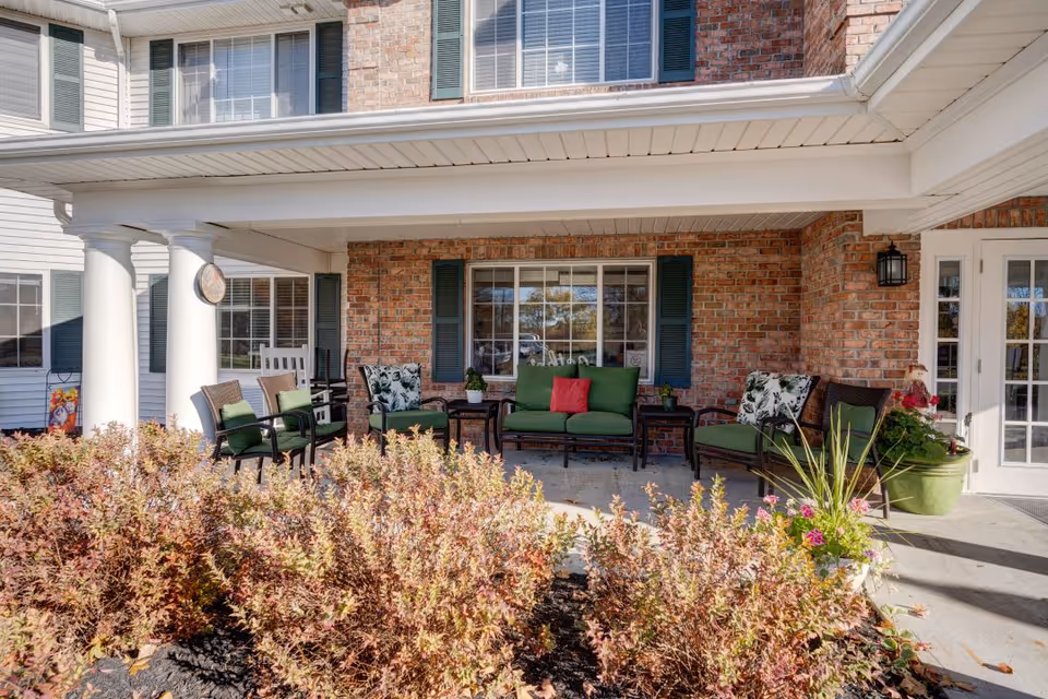 Covered outdoor patio area with brick walls and white columns, furnished with green cushioned chairs and a loveseat, a small table, and surrounded by potted plants and shrubs in front.