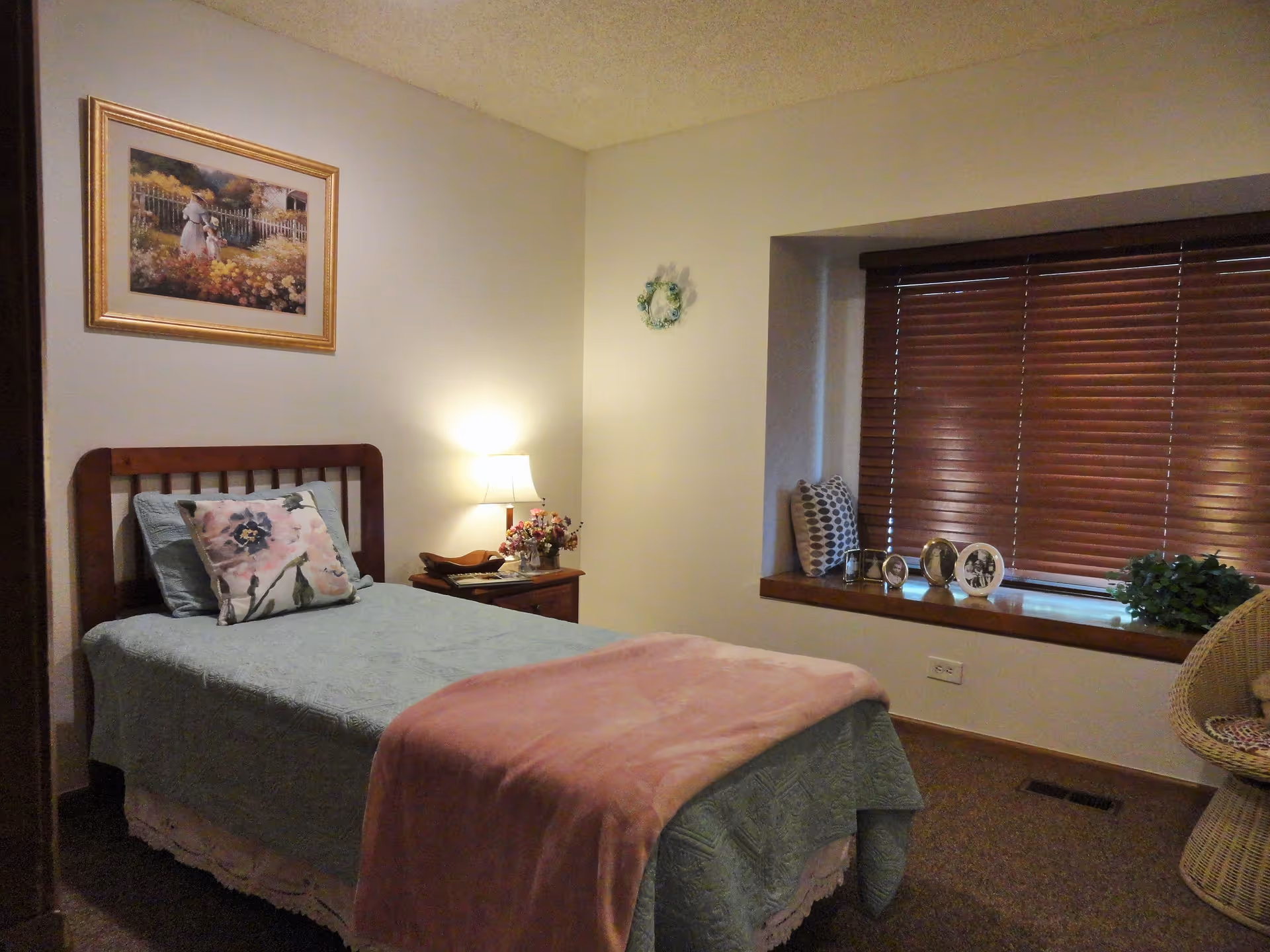 A cozy bedroom with a single wooden bed covered with a light blue bedspread and a pink throw blanket. There is a floral pillow on the bed. Next to the bed is a wooden nightstand with a lamp and a small flower arrangement. A framed painting hangs on the wall above the bed. The room has a window with wooden blinds and a cushioned window seat decorated with pillows and framed photos. A wicker chair is partially visible on the right side.
