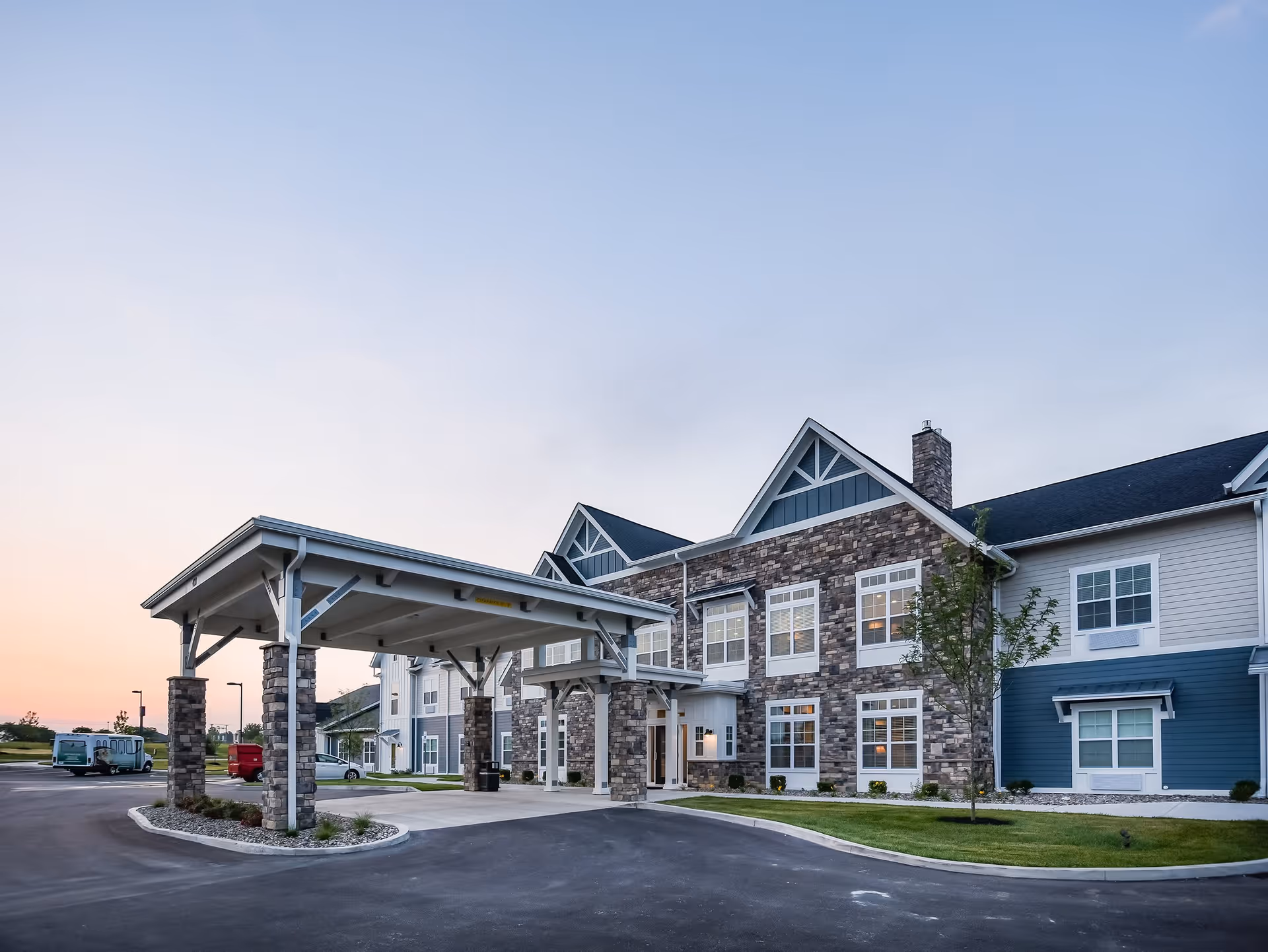 Exterior view of Traditions at Hunter Station, a senior living facility with a covered entrance supported by stone pillars, a stone and siding facade, multiple windows, and a paved driveway with parked vehicles in the background during early evening.