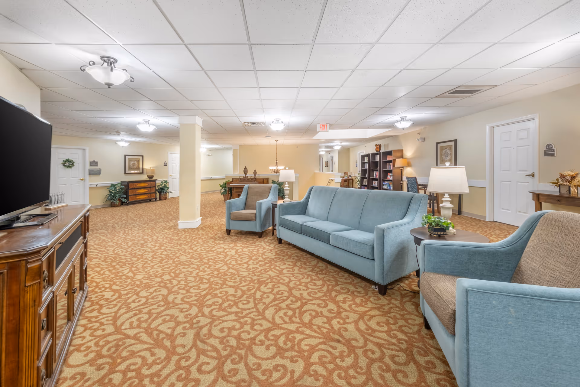 A spacious senior living facility common area with patterned carpet, light blue upholstered sofa and armchairs, wooden TV stand with a large flat-screen TV, lamps, plants, bookshelves, and framed artwork on the walls under a white tiled ceiling with multiple light fixtures.