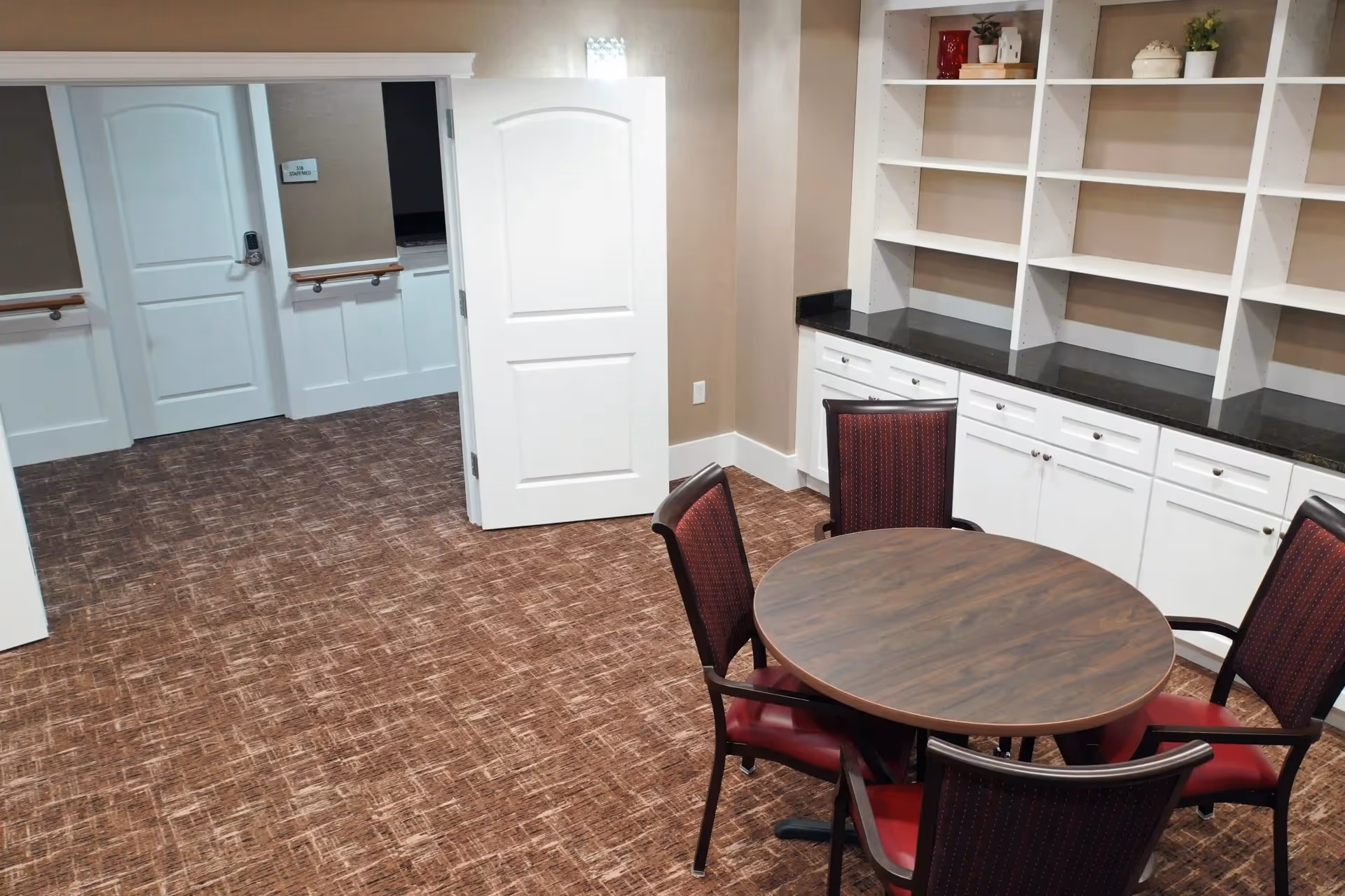 Interior room with a round wooden table surrounded by four chairs with red cushions. The room has brown patterned carpet, beige walls, white baseboards, and white built-in shelves and cabinets with a black countertop. Two white doors are open, leading to a hallway with similar decor.