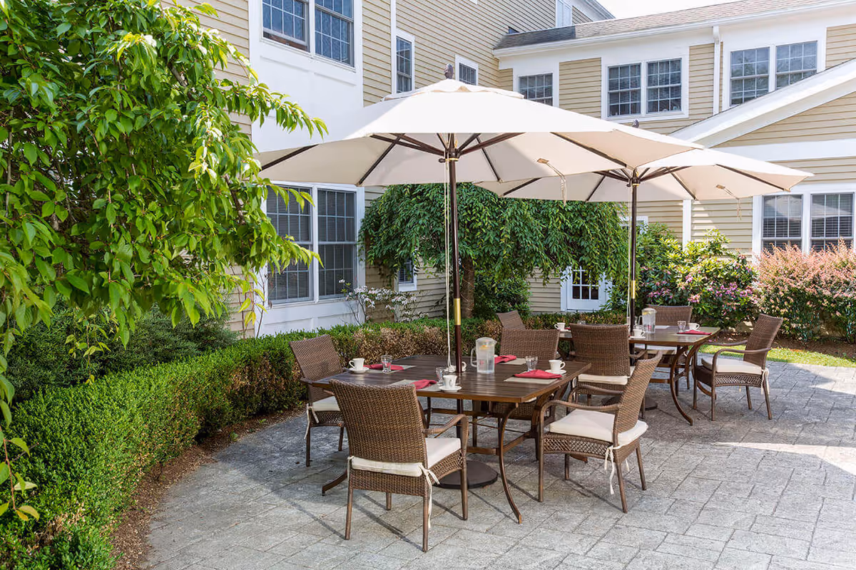Outdoor patio area at Benchmark at Ridgefield Crossings with two tables, each shaded by large white umbrellas. The tables are set with cups, glasses, and napkins. Surrounding the patio are green bushes, trees, and the exterior walls of the building with multiple windows.