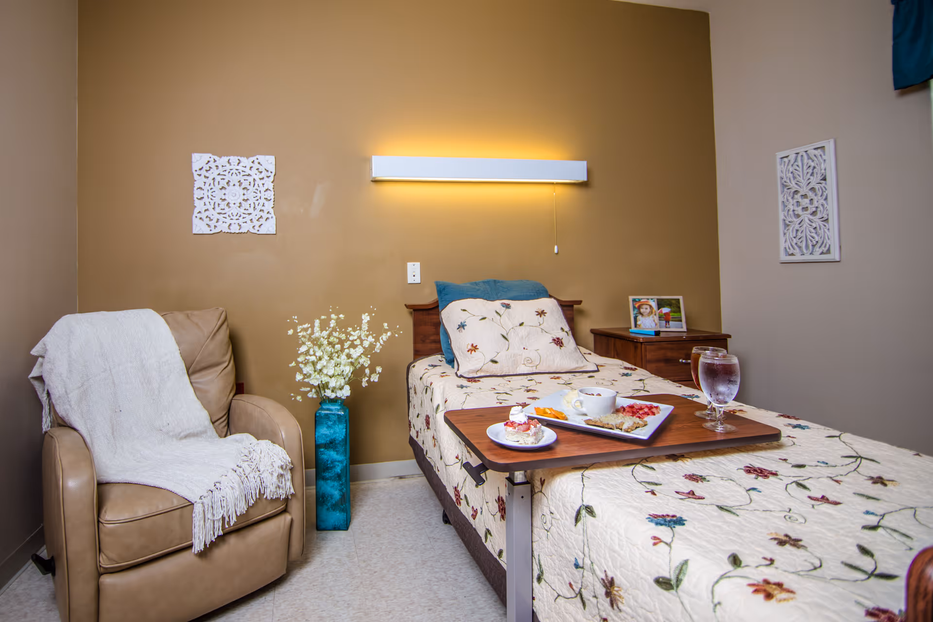 A neatly decorated resident bedroom with a single bed, overbed meal tray, armchair, and wall-mounted light.