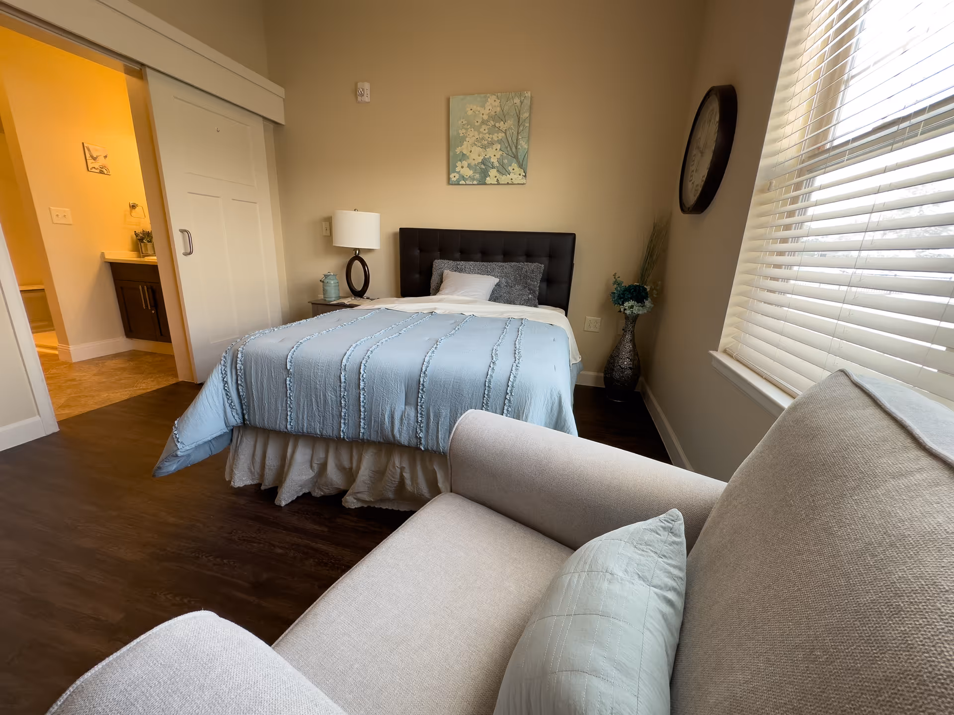 A cozy bedroom with a bed covered in a light blue quilt and pillows, a bedside table with a lamp and decorative items, a large window with blinds, a beige armchair with a matching pillow, a wall clock, and a floral painting above the bed. A doorway leads to a bathroom area with a sink and mirror visible.