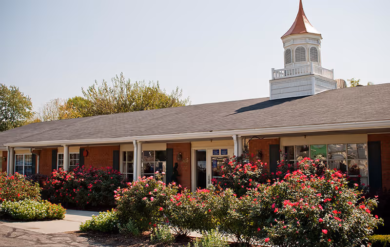 Exterior view of a single-story brick building with a gray shingled roof and a white cupola on top. The front of the building is lined with green bushes and vibrant pink flowers. There is a sidewalk leading to the entrance, and trees are visible in the background under a clear sky.
