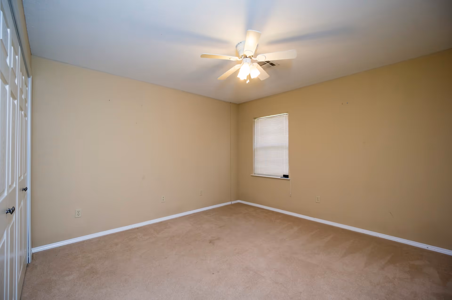 Empty room with beige walls and carpet, a ceiling fan with lights, a window with closed blinds, and a set of white double doors on the left side.
