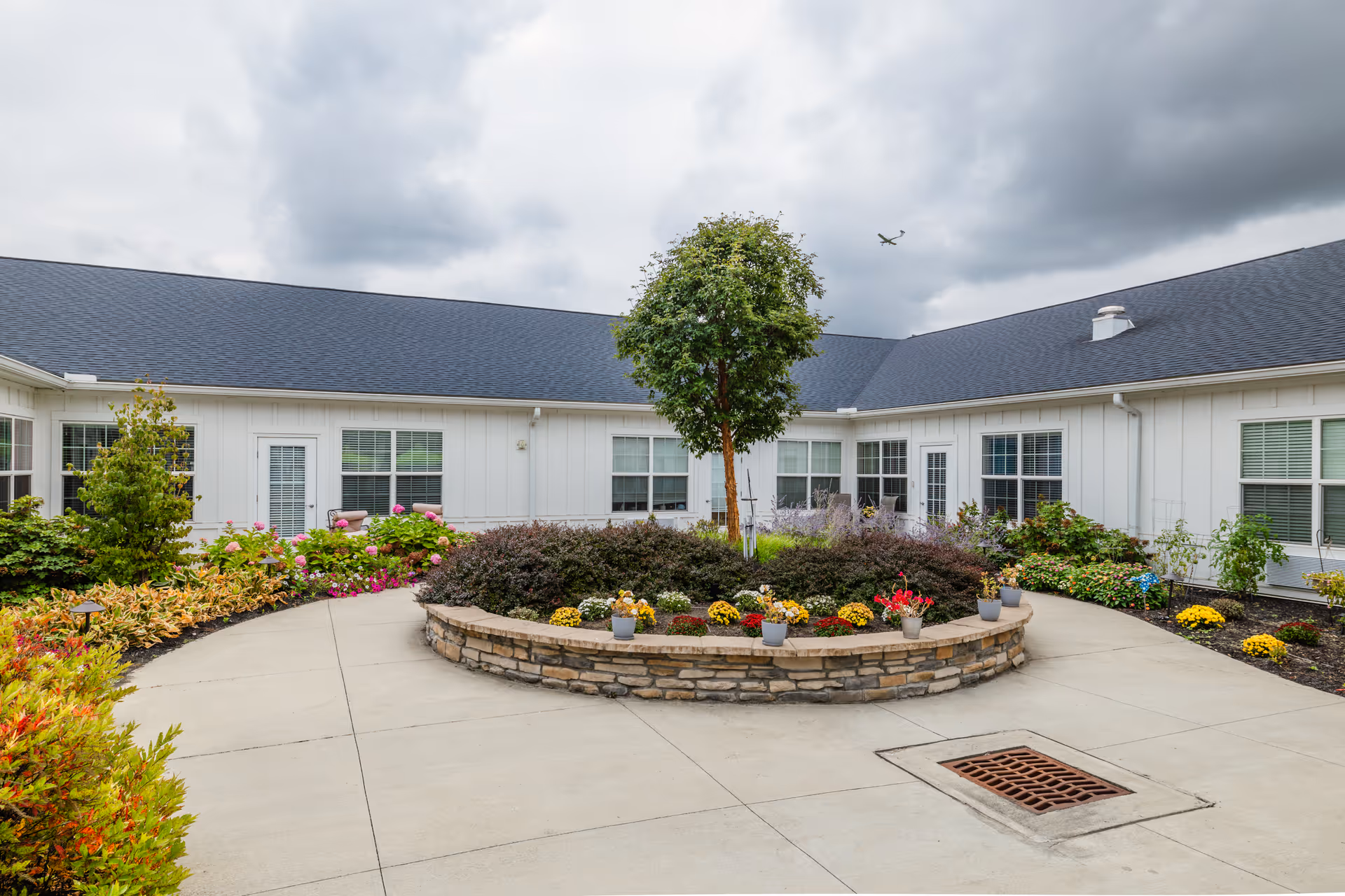 Outdoor courtyard area at Walnut Crossing featuring a circular raised stone planter with a small tree and various colorful flowers. The courtyard is surrounded by a white building with multiple windows and doors, under a cloudy sky with an airplane visible in the distance.