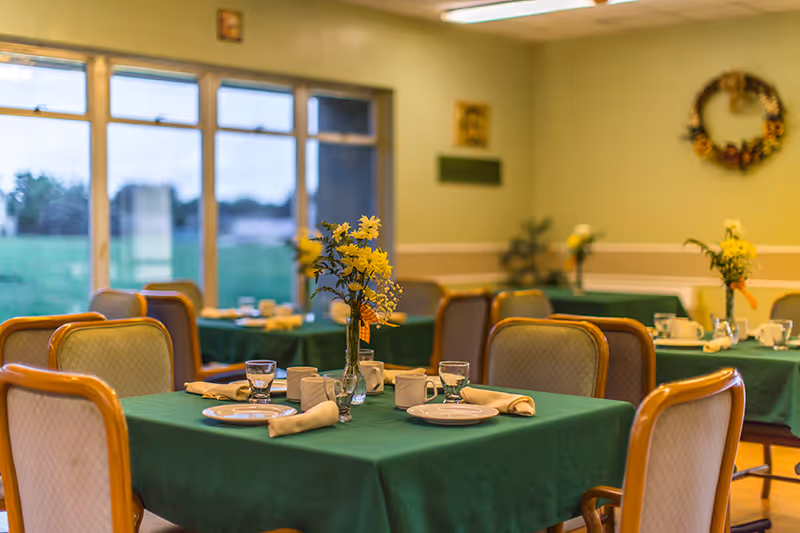 A dining room with tables covered in green tablecloths, each set with plates, cups, glasses, and napkins. Yellow flower centerpieces are placed on each table. The room has large windows letting in natural light and light green walls decorated with a wreath and framed pictures.
