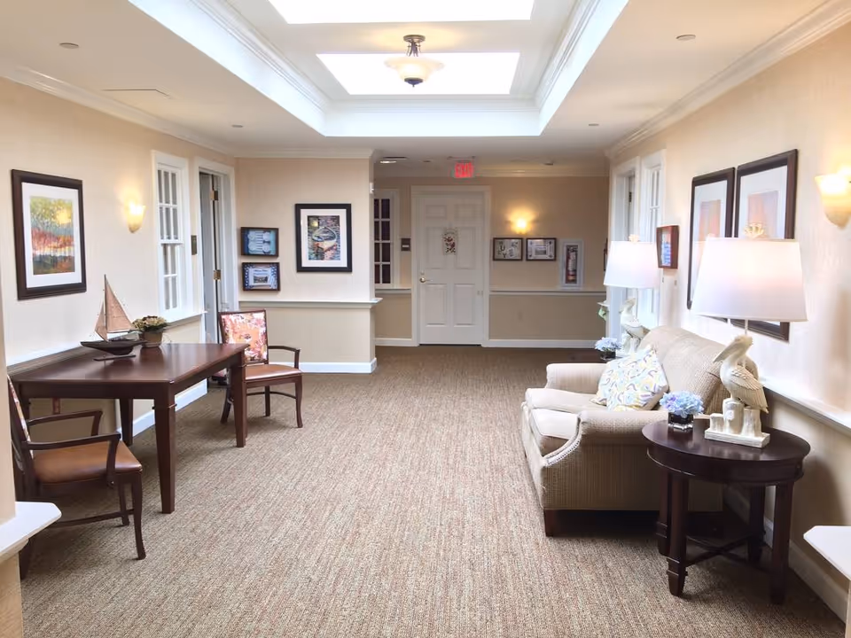 A well-lit hallway in a senior living facility with beige walls and carpeted floor. On the left side, there is a wooden table with two chairs and a decorative sailboat on top. On the right side, there is a beige upholstered sofa with patterned pillows, a round wooden side table with a lamp and decorative items, and framed artwork on the walls. The hallway leads to a white door at the end, with additional framed pictures on the walls.