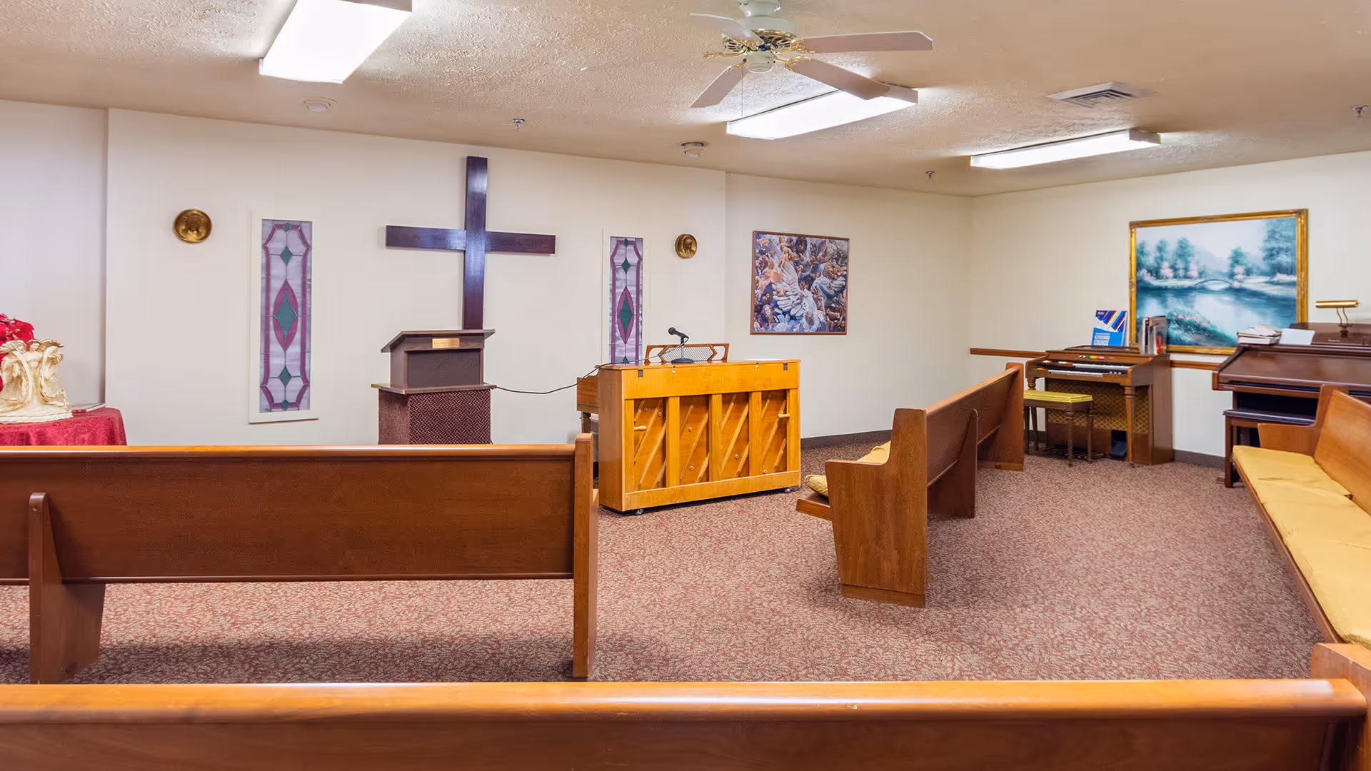 Interior view of a small chapel or worship room with wooden pews, a wooden cross on the wall, a podium, a piano, and religious artwork. The room has carpeted floors, ceiling fans, and soft lighting.