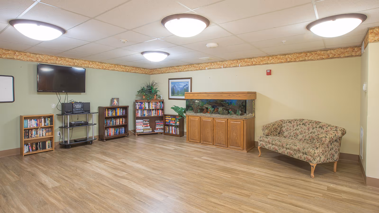 A cozy common area with wooden flooring, three ceiling lights, a floral-patterned couch, a large fish tank on a wooden cabinet, several bookshelves filled with books, a flat-screen TV mounted on the wall, and a stereo system on a metal stand. The walls are painted light green and beige with a decorative border near the ceiling.
