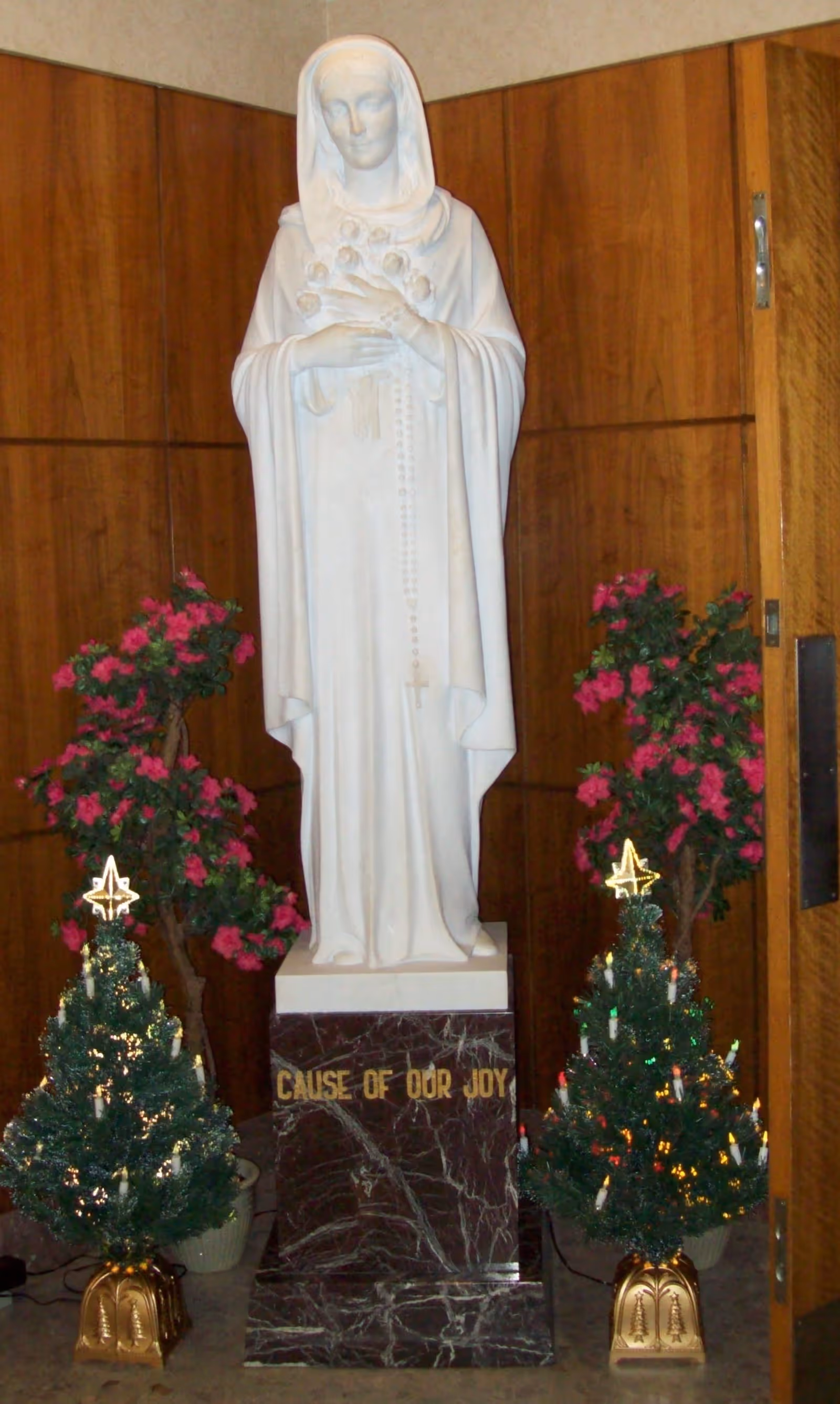 A white statue of a woman holding roses and a rosary, standing on a dark marble pedestal with the inscription 'CAUSE OF OUR JOY'. The statue is flanked by two small decorated Christmas trees with lights and stars on top, and two potted plants with pink flowers. The background features wood-paneled walls.
