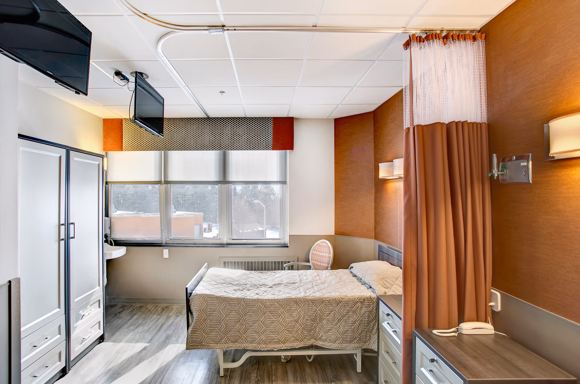 A bright and clean rehabilitation center bedroom with a single bed covered in a beige patterned bedspread. The room features a large window with blinds, a wall-mounted TV, a wardrobe, a chair, a small sink, and a privacy curtain. The walls are decorated with warm brown tones and modern light fixtures.