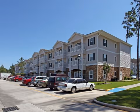 Exterior view of a three-story residential building with balconies, surrounded by a parking lot with several parked cars under a clear blue sky.