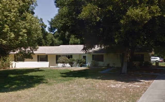 Single-story building with a light-colored exterior surrounded by trees and a grassy lawn, with a sidewalk leading up to the entrance.