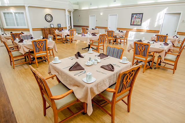 A dining room in a senior living facility with multiple tables covered in beige tablecloths, each set with white cups, saucers, silverware, and folded brown napkins. Wooden chairs with cushioned seats surround the tables. The room has light-colored wooden flooring, white walls, and several doors and framed artwork on the walls.