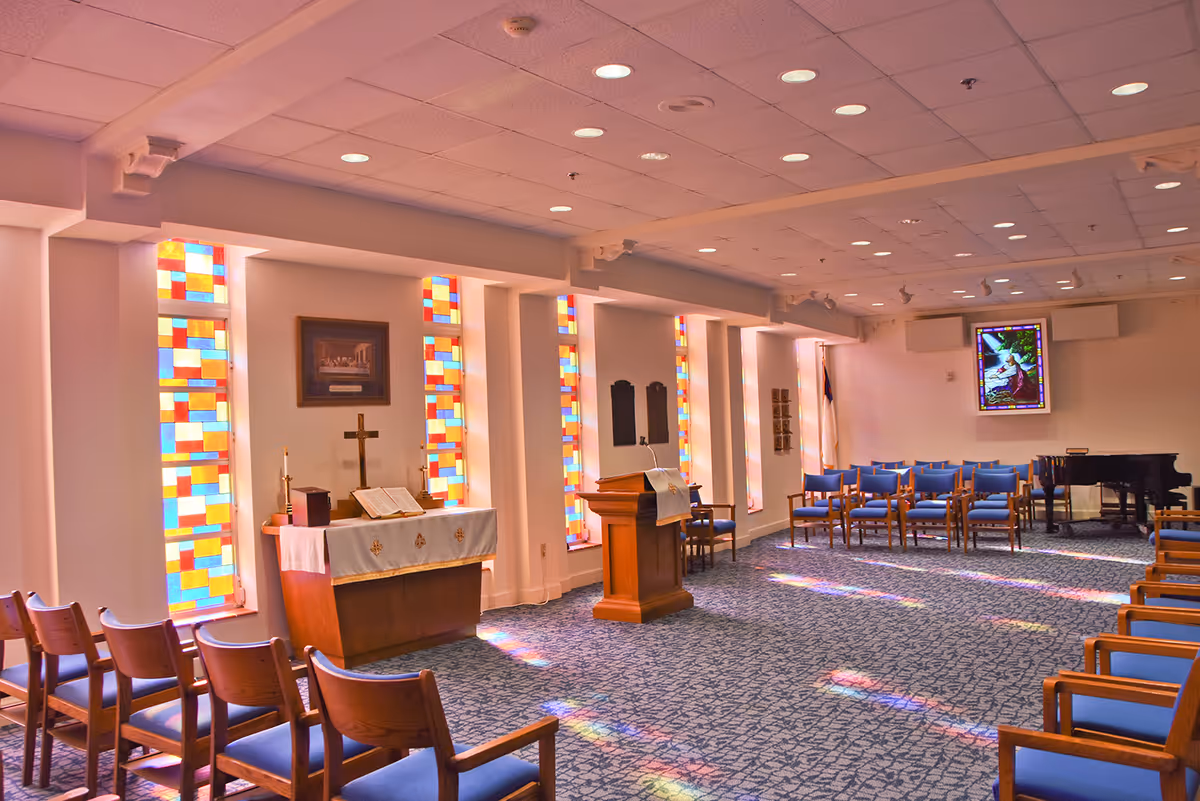 Interior of a chapel or worship room with rows of wooden chairs with blue cushions, stained glass windows casting colorful light patterns on the carpeted floor, a wooden altar with a cross and open Bible, a lectern with a cloth, and a grand piano in the corner.
