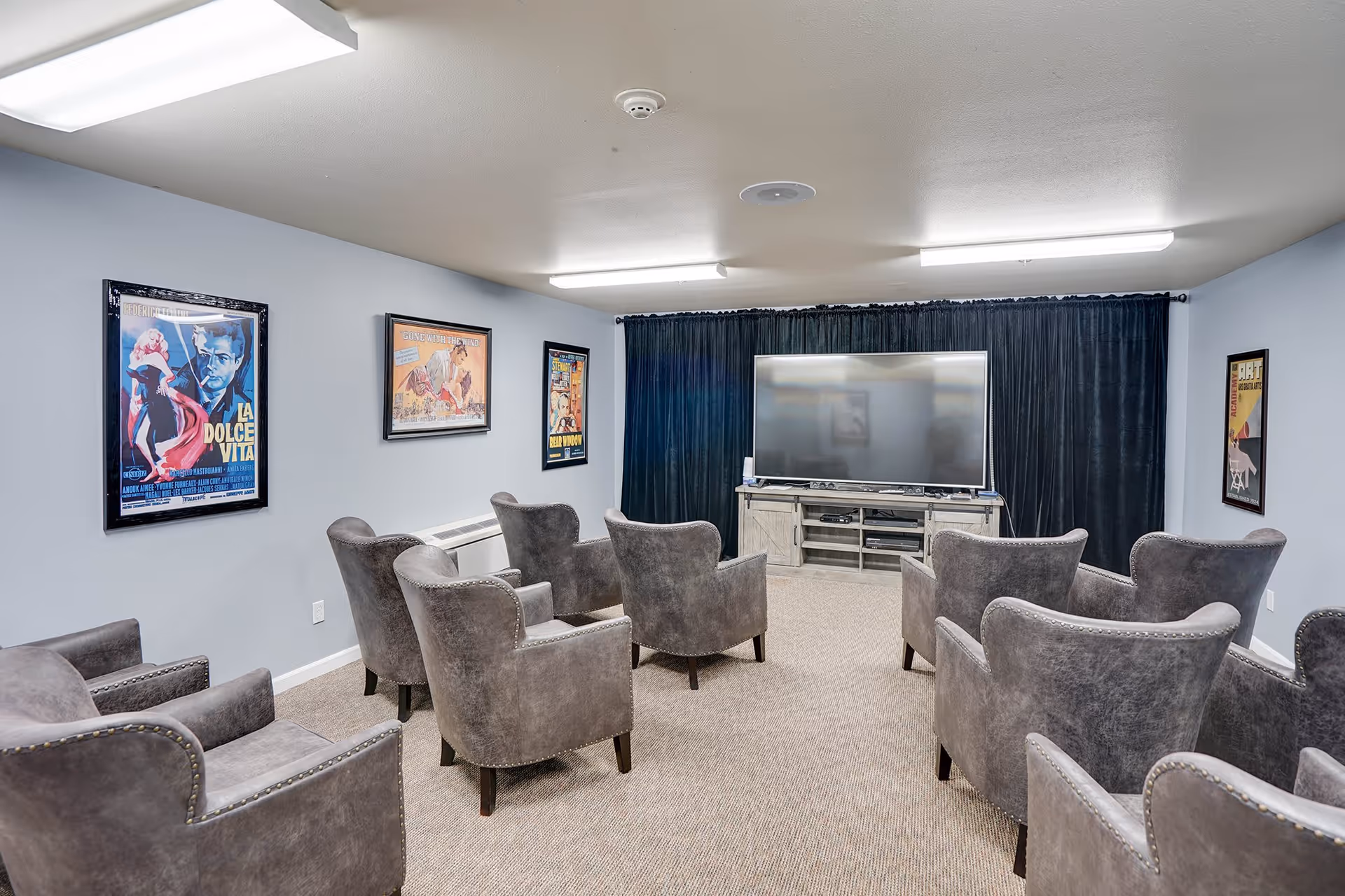 A small theater-style room with multiple gray upholstered armchairs arranged in rows facing a large flat-screen TV mounted on a wooden media console. The walls are light blue and decorated with framed vintage movie posters. The room has a carpeted floor and fluorescent ceiling lights.