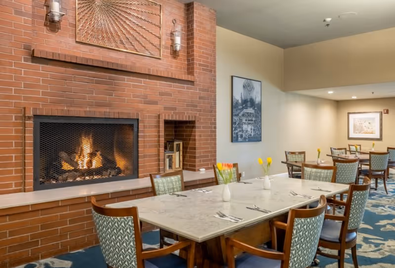 A dining area with a long marble table set with utensils and small vases holding yellow and orange flowers. The room features a brick fireplace with a fire burning inside, wall-mounted lights, framed artwork on the walls, and several wooden chairs with patterned upholstery. The carpet has a blue and beige floral design.