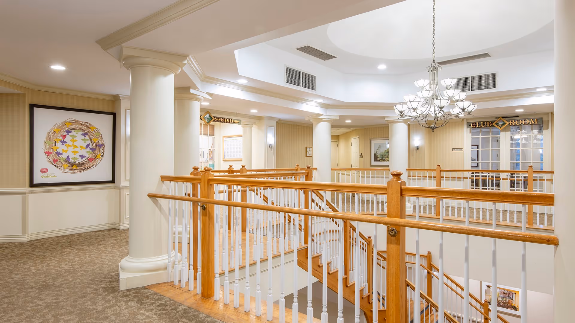 Interior view of a senior living facility hallway with a wooden handrail staircase in the center, white columns, beige walls, carpeted floors, and a chandelier hanging from the ceiling. There are signs indicating a studio and a club room in the background, along with framed artwork on the walls.