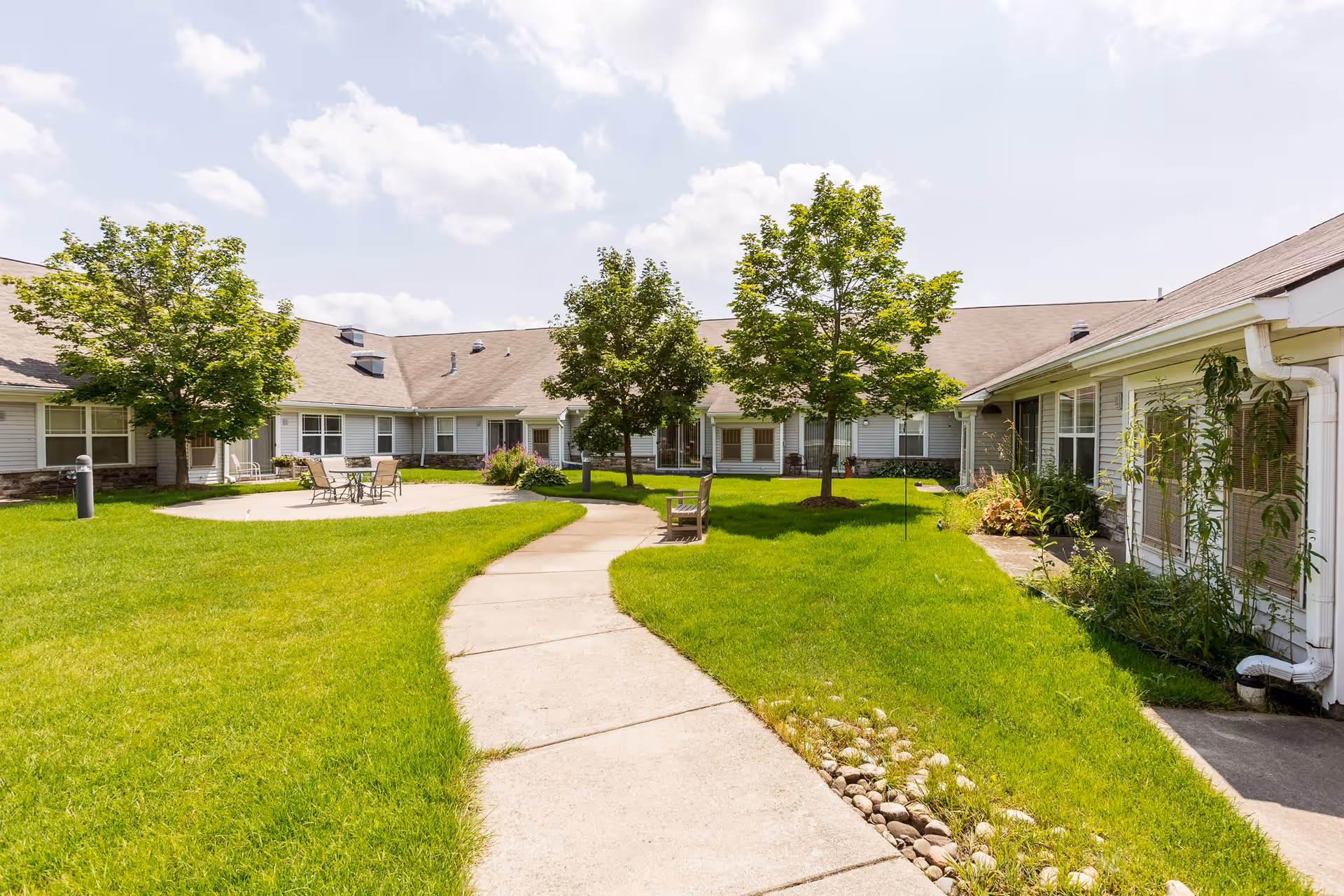 A sunny outdoor courtyard area at Independence Village of Oxford (Waterstone) featuring a concrete walkway, green grass, several trees, a few benches, and a round patio with a table and chairs. The courtyard is surrounded by single-story building structures with windows and light-colored siding.