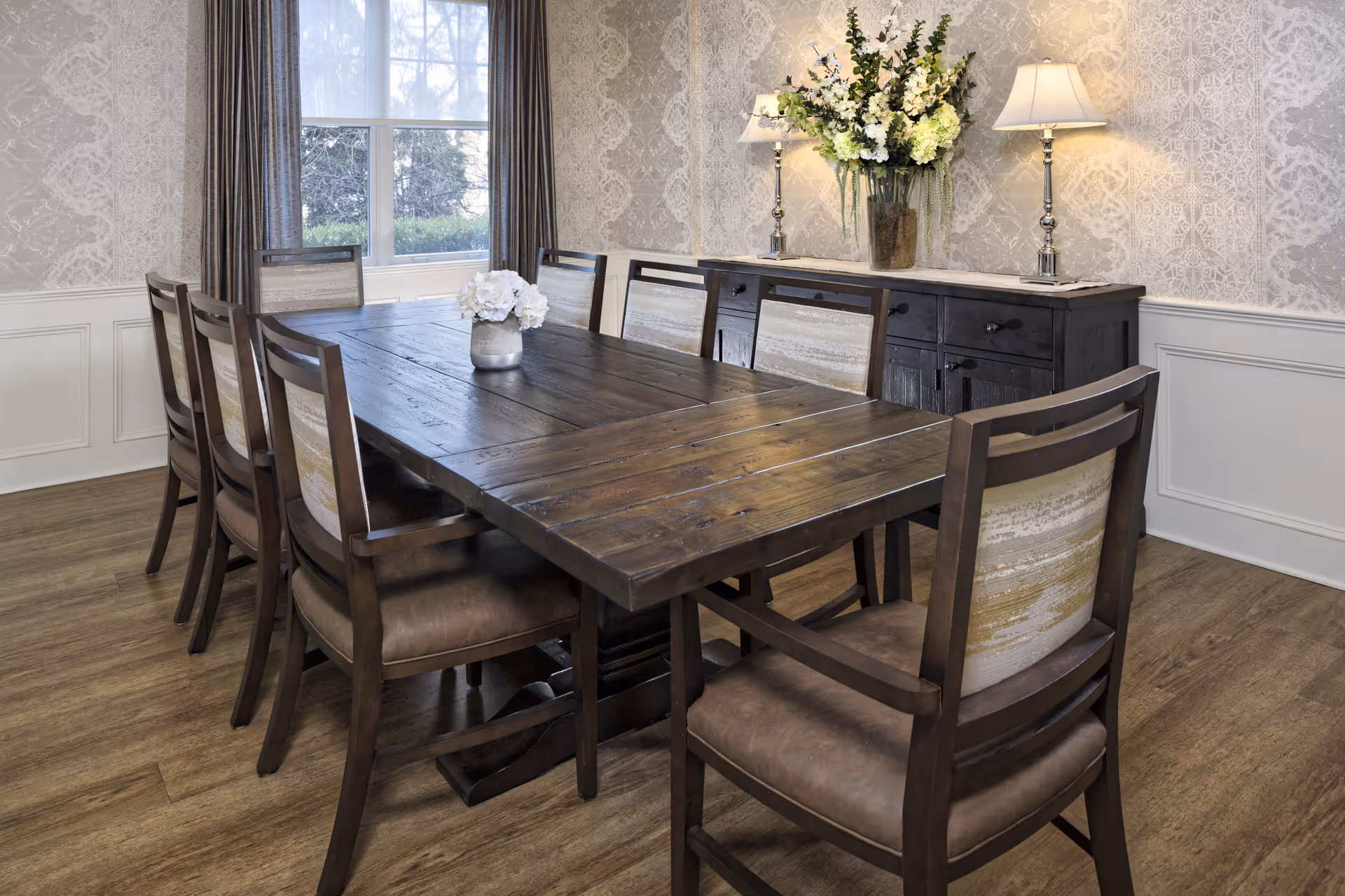 A dining room with a long wooden table surrounded by eight chairs with cushioned seats and backs. A window with curtains is visible in the background, along with a dark wooden sideboard topped with two lamps and a large vase of flowers. The walls have a patterned wallpaper and white wainscoting, and the floor is wooden.