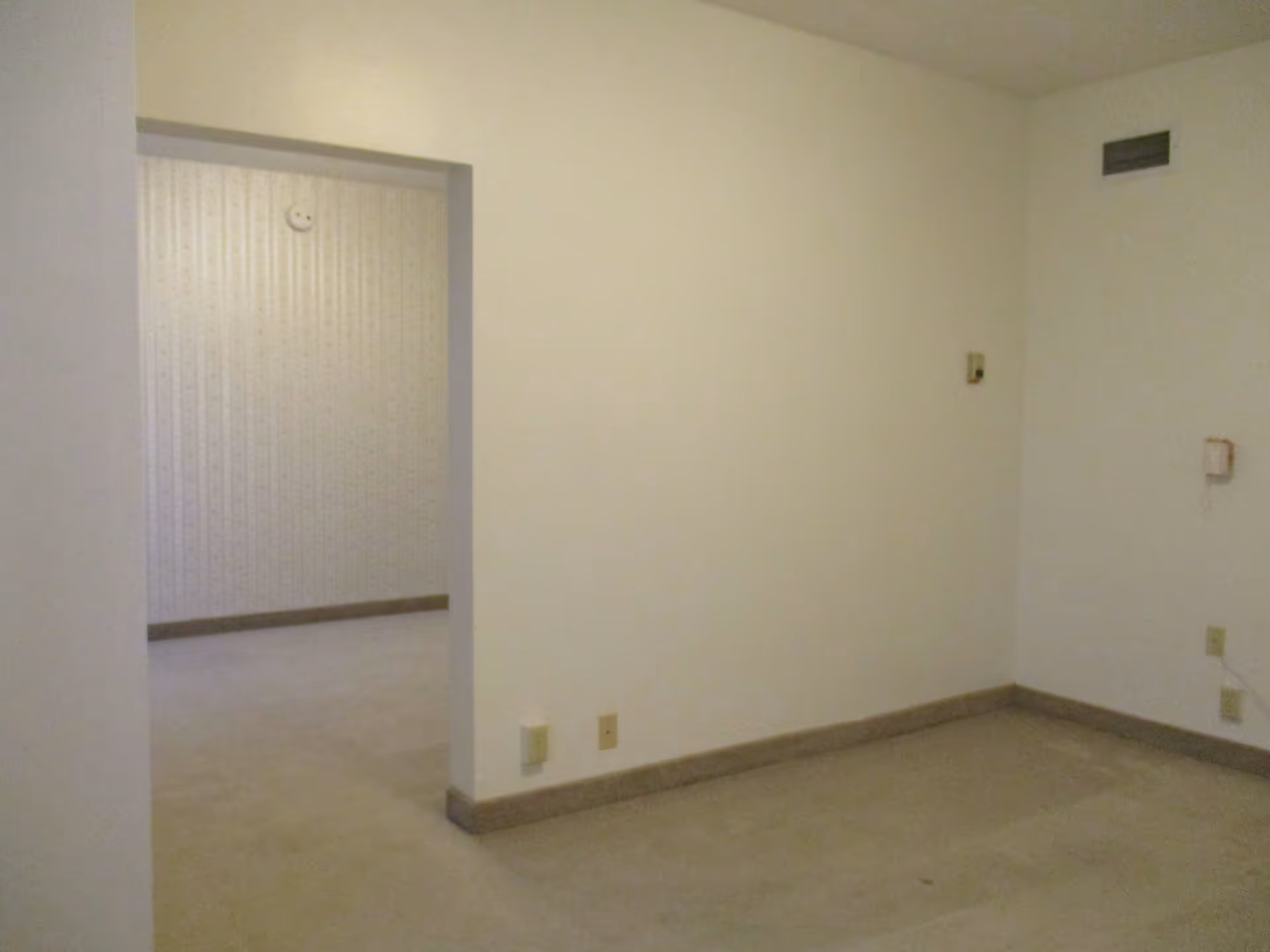 An empty interior room with beige carpet and off-white walls. There is a doorway leading to another room with patterned wallpaper. The room has baseboards and some electrical outlets and switches visible on the walls.