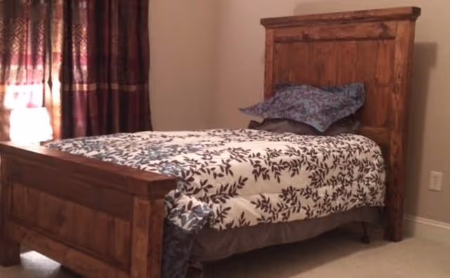 A bedroom with a wooden bed frame featuring a headboard and footboard. The bed is made with a white comforter decorated with a dark leaf pattern and has two pillows, one blue and one gray. There is a window with dark patterned curtains and a lit lamp on a nightstand beside the bed.