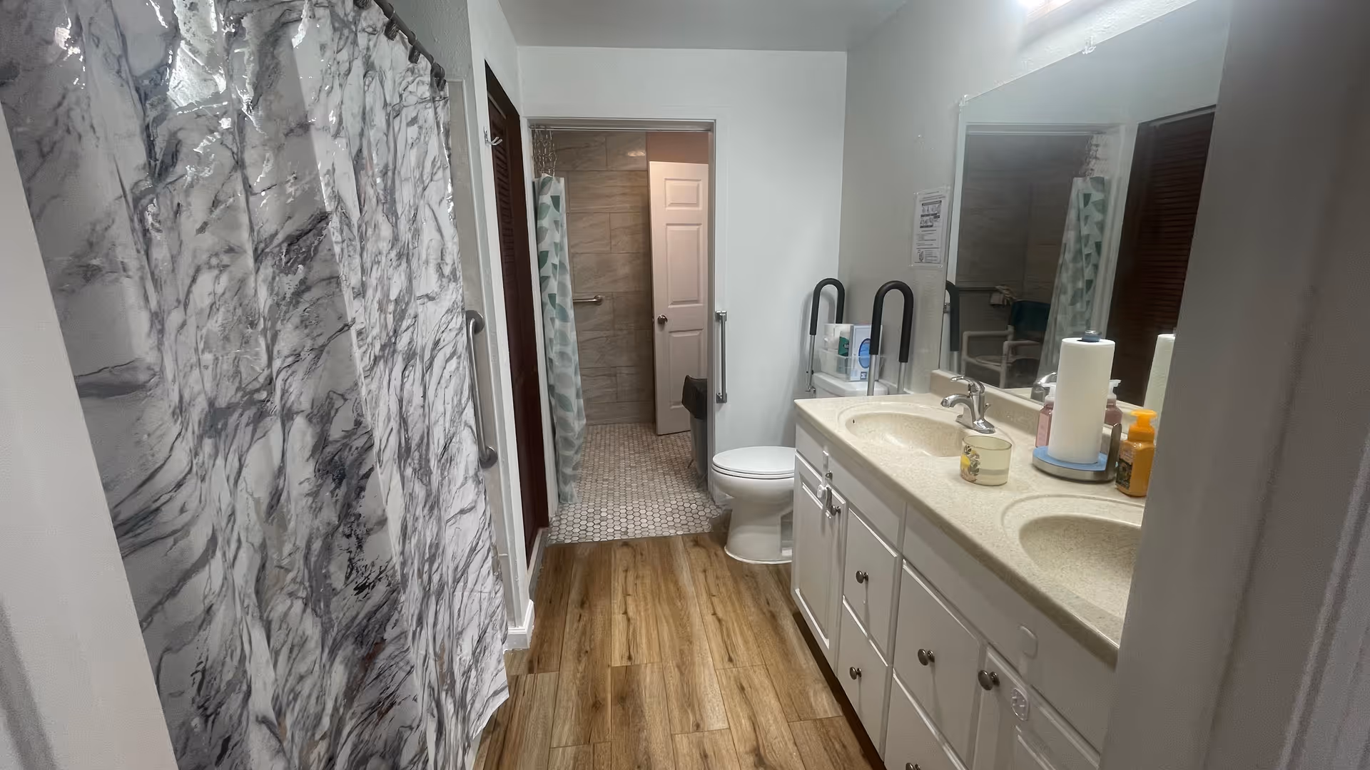 A bathroom with a double sink vanity on the right, a toilet next to it, and a shower with a marble-patterned curtain on the left. The floor is wood-style laminate, and there is a doorway leading to another room with a tiled floor and a second shower with a patterned curtain.