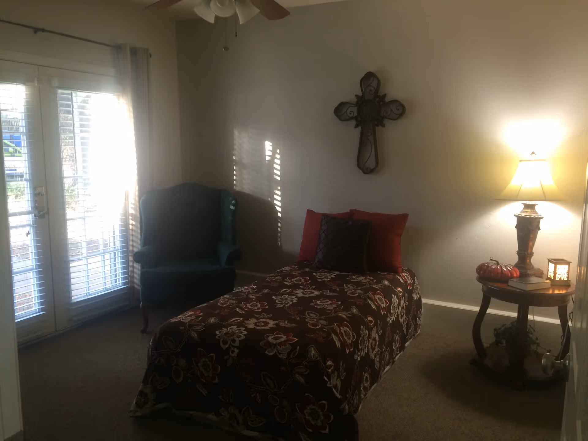 Small bedroom with a single bed in floral bedding, an armchair by glass doors, and a side table with a lamp.