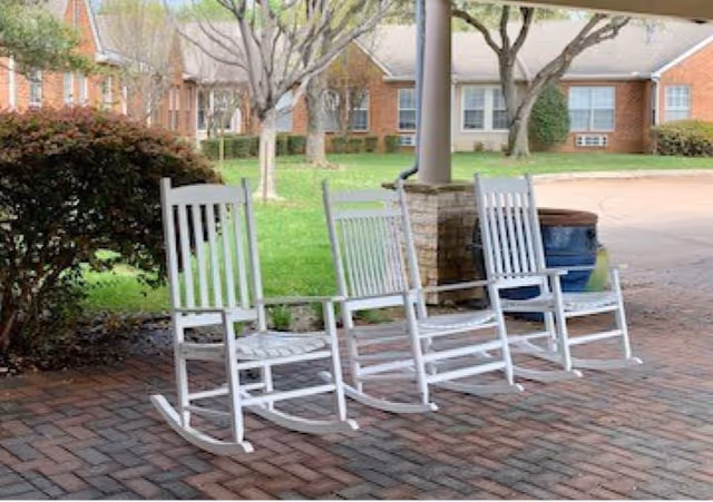 Three white wooden rocking chairs on a covered brick patio overlooking a lawn with trees and brick residential buildings.