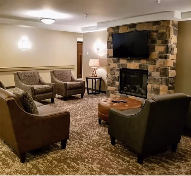 A cozy living room area with a stone fireplace and a flat-screen TV mounted above it. The room features four upholstered armchairs arranged around a round leather ottoman. A small side table with a lamp is placed next to the fireplace. The walls are painted in a light neutral color and the floor is carpeted with a patterned design.