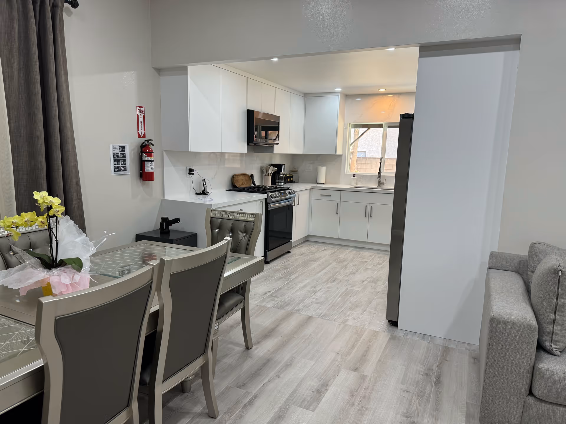 Open-plan modern kitchen and dining area with white cabinets, stainless appliances, a dining table with chairs and a gray sofa at the edge.