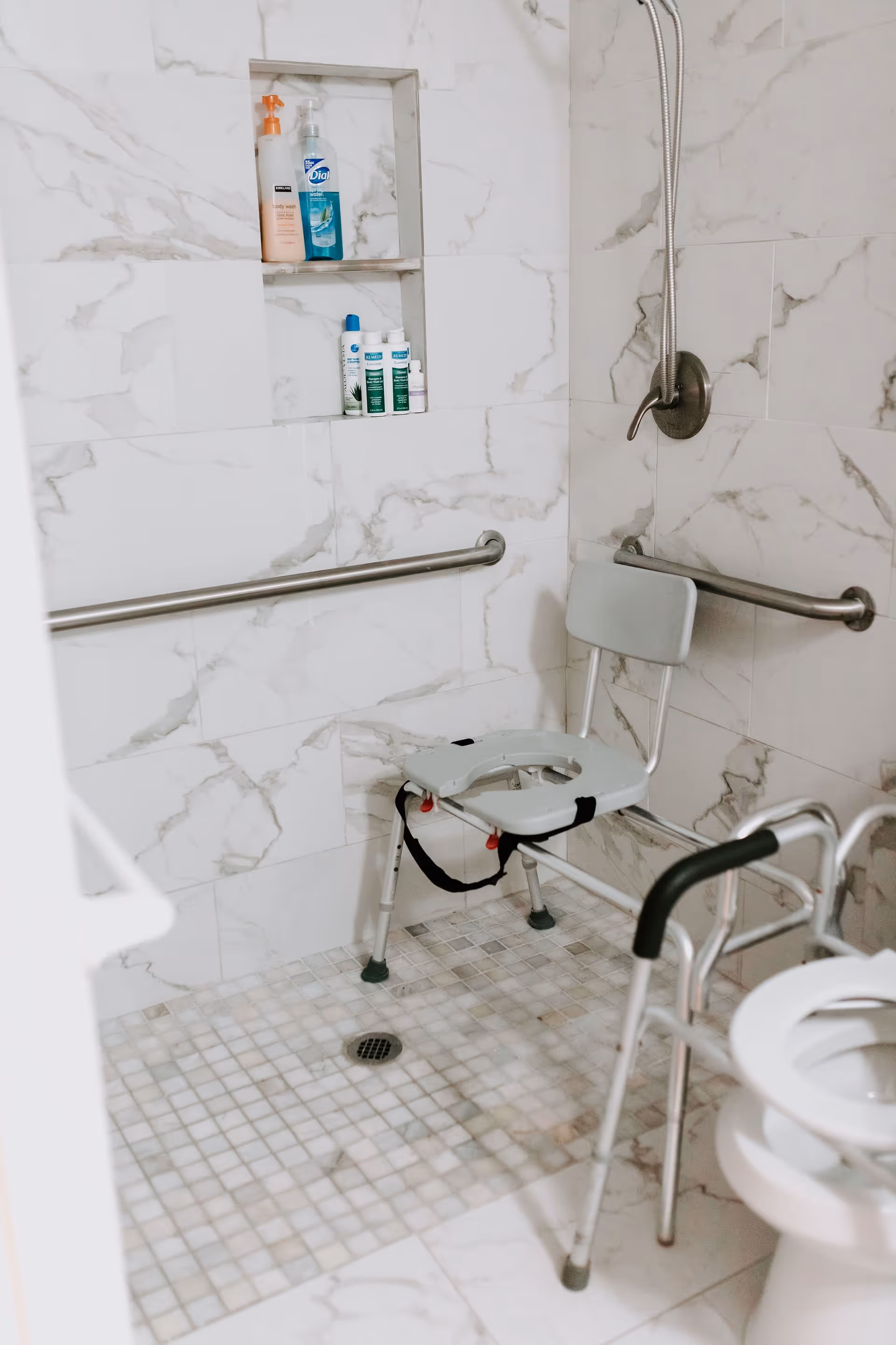 Accessible shower area with marble-patterned tiles, a shower chair with a backrest and safety straps, grab bars on the walls, built-in shelves holding toiletries, and a partial view of a walker and toilet seat.
