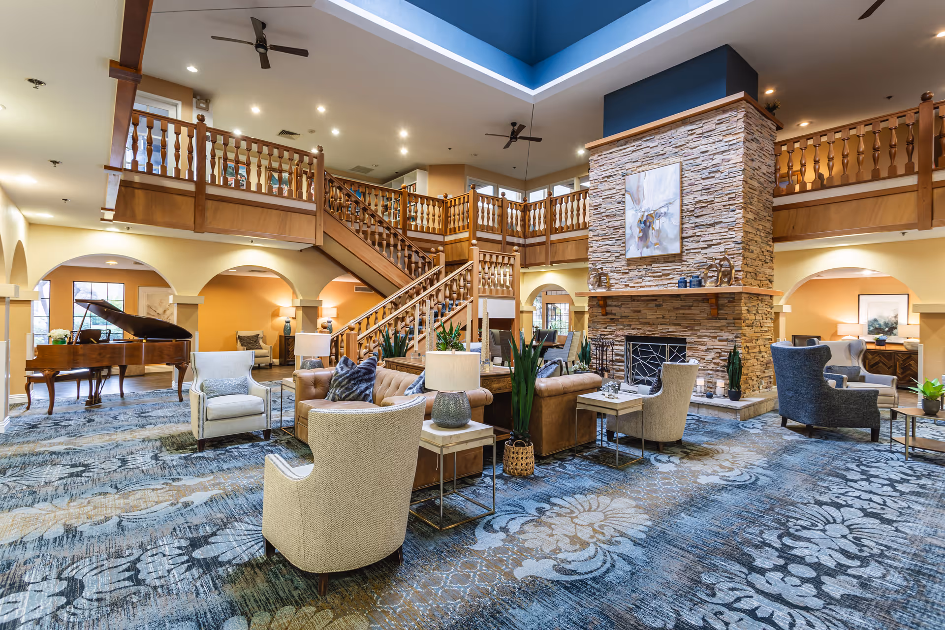 Spacious senior living facility common area with a large stone fireplace, comfortable seating including armchairs and sofas, a grand piano, and a wooden staircase leading to an upper balcony. The room features high ceilings with ceiling fans and warm lighting.