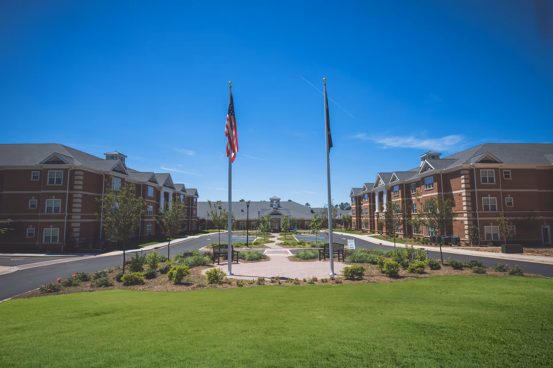 Front exterior of a senior living campus with two three-story brick buildings flanking a landscaped courtyard and flagpoles.