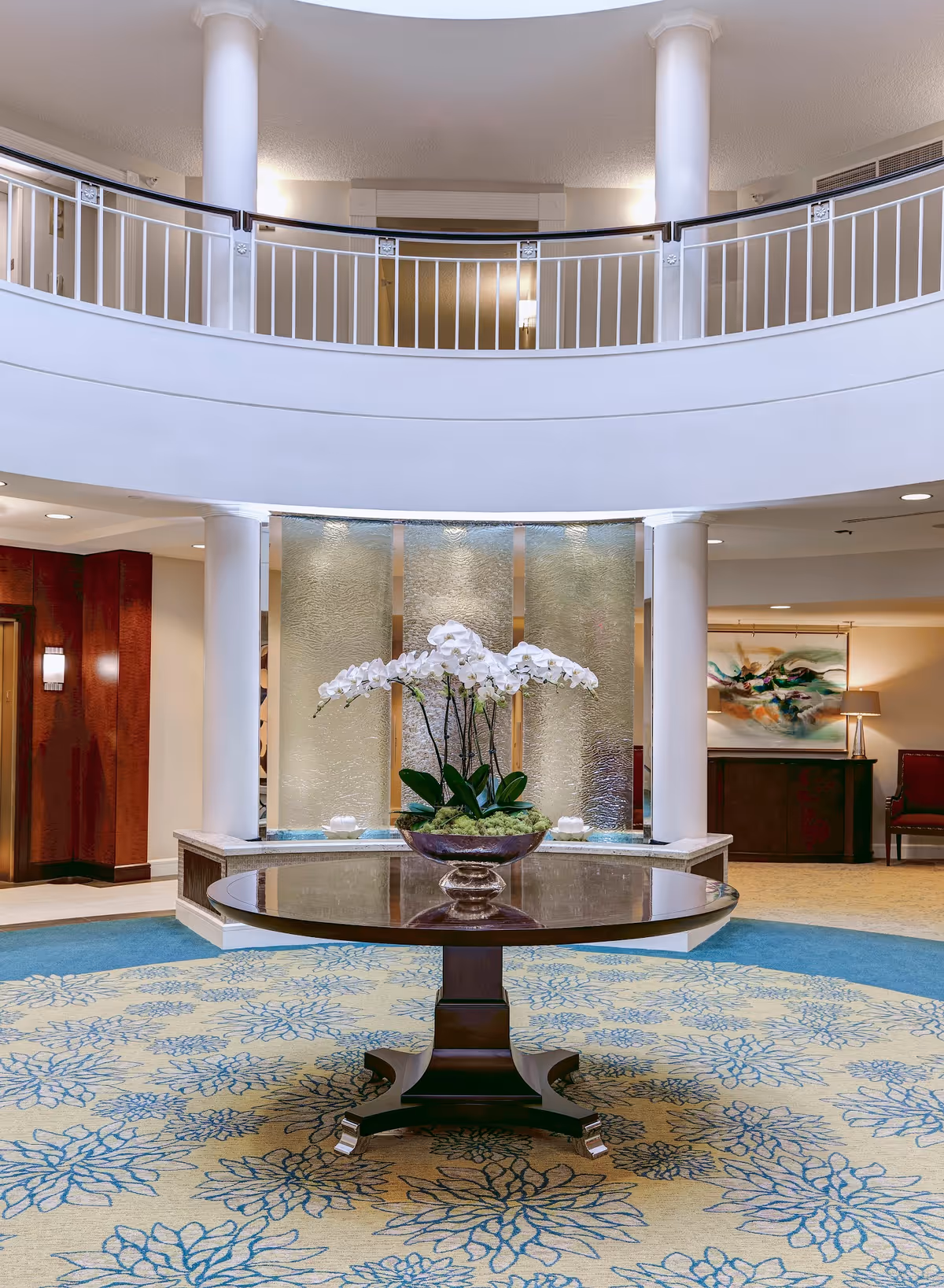 A spacious and elegant lobby area with a round wooden table in the center holding a large vase of white orchids. The floor is covered with a patterned carpet in blue and beige tones. Behind the table is a decorative water feature with textured glass panels, flanked by white columns. The upper level has a white railing overlooking the lobby. The walls are adorned with artwork and soft lighting.