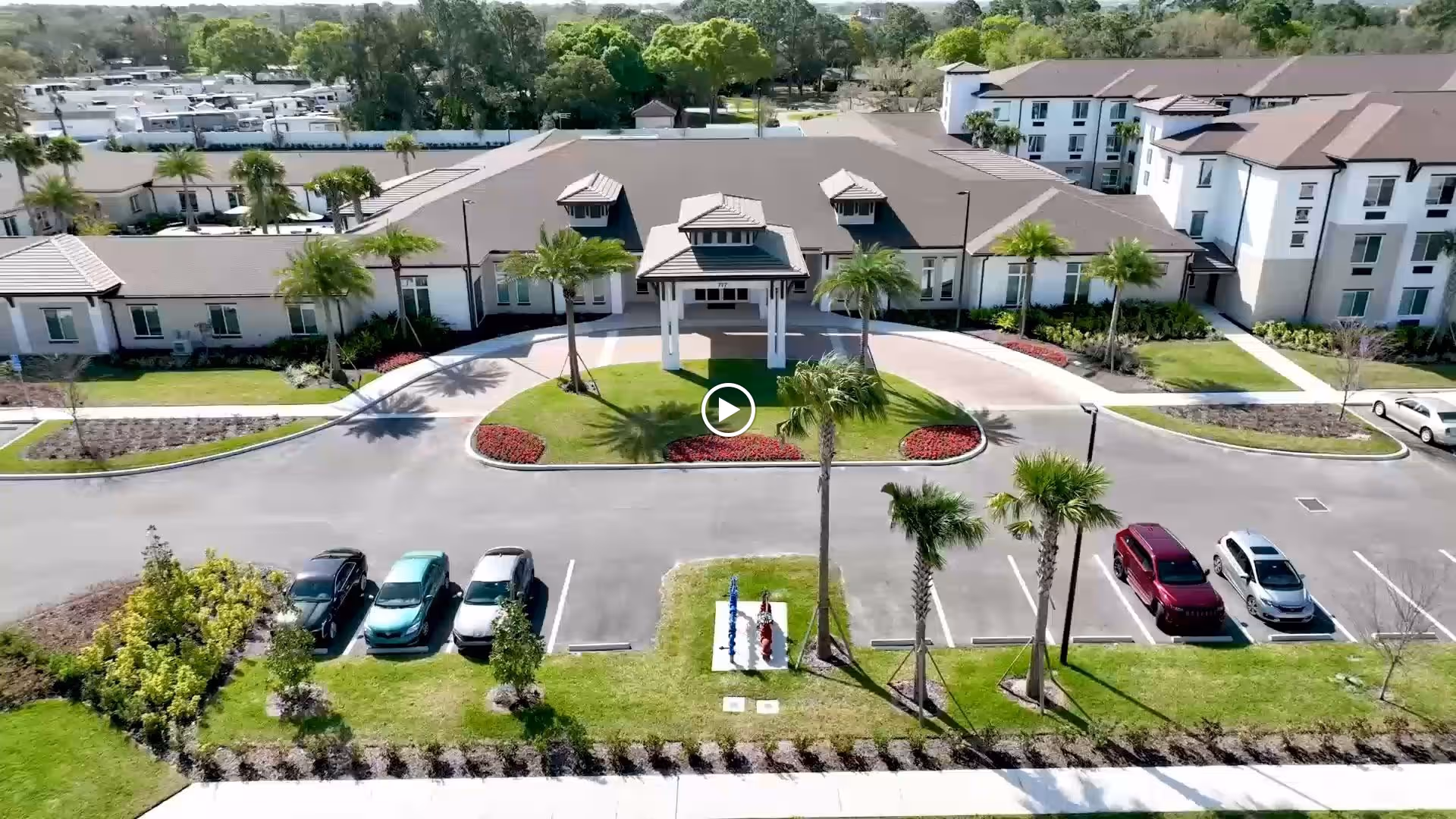 Aerial view of a senior living facility named Alura Senior Living showing a large building with a covered entrance, surrounded by palm trees, landscaped gardens, and a parking lot with several cars parked.
