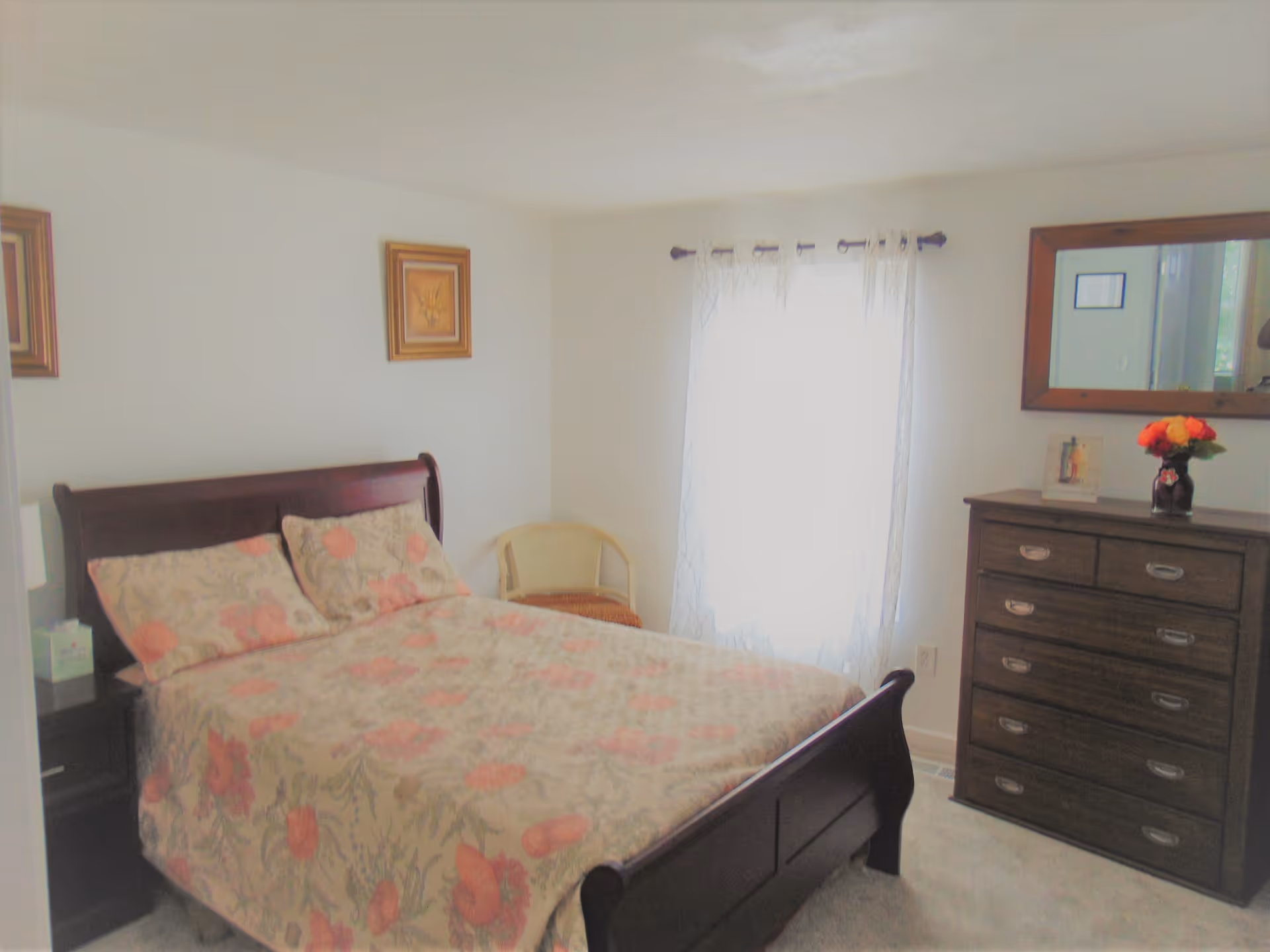 Sunlit bedroom with a wooden sleigh bed covered in a floral bedspread, a dresser with mirror and a window with sheer curtains.