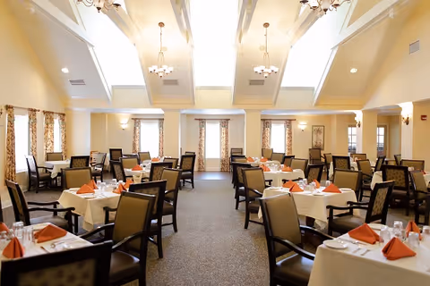 Bright, elegant dining room with multiple tables set with white tablecloths, orange napkins, and upholstered chairs under skylights.