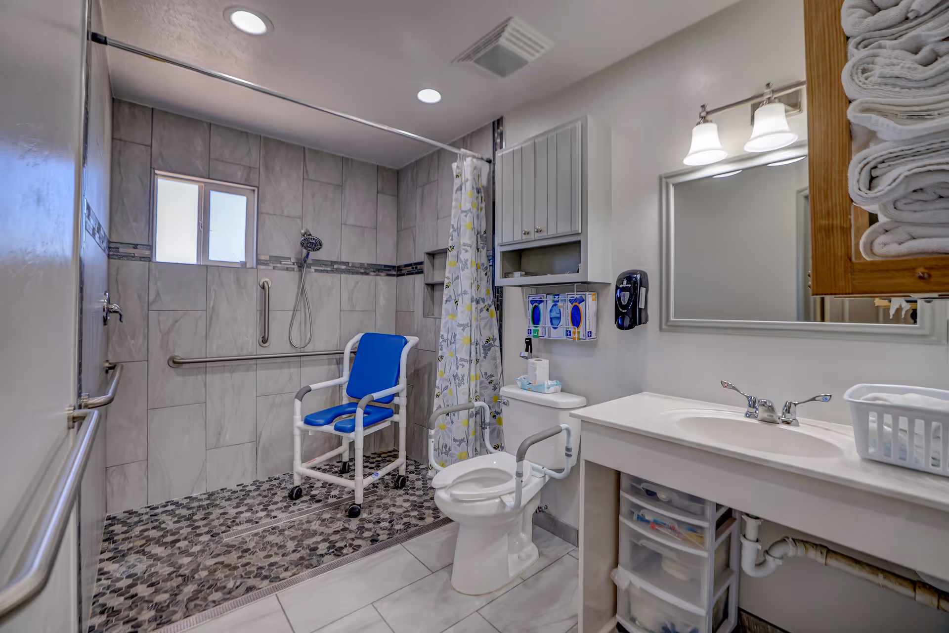 Accessible bathroom with a roll-in tiled shower, blue shower chair, grab bars, toilet with armrests, sink and mirror with stacked towels.