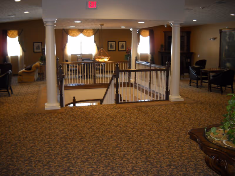 Interior view of a senior living facility common area with patterned carpet, white columns, a staircase with wooden railing, and seating areas with chairs and sofas near windows with curtains. A chandelier hangs above the stairwell.